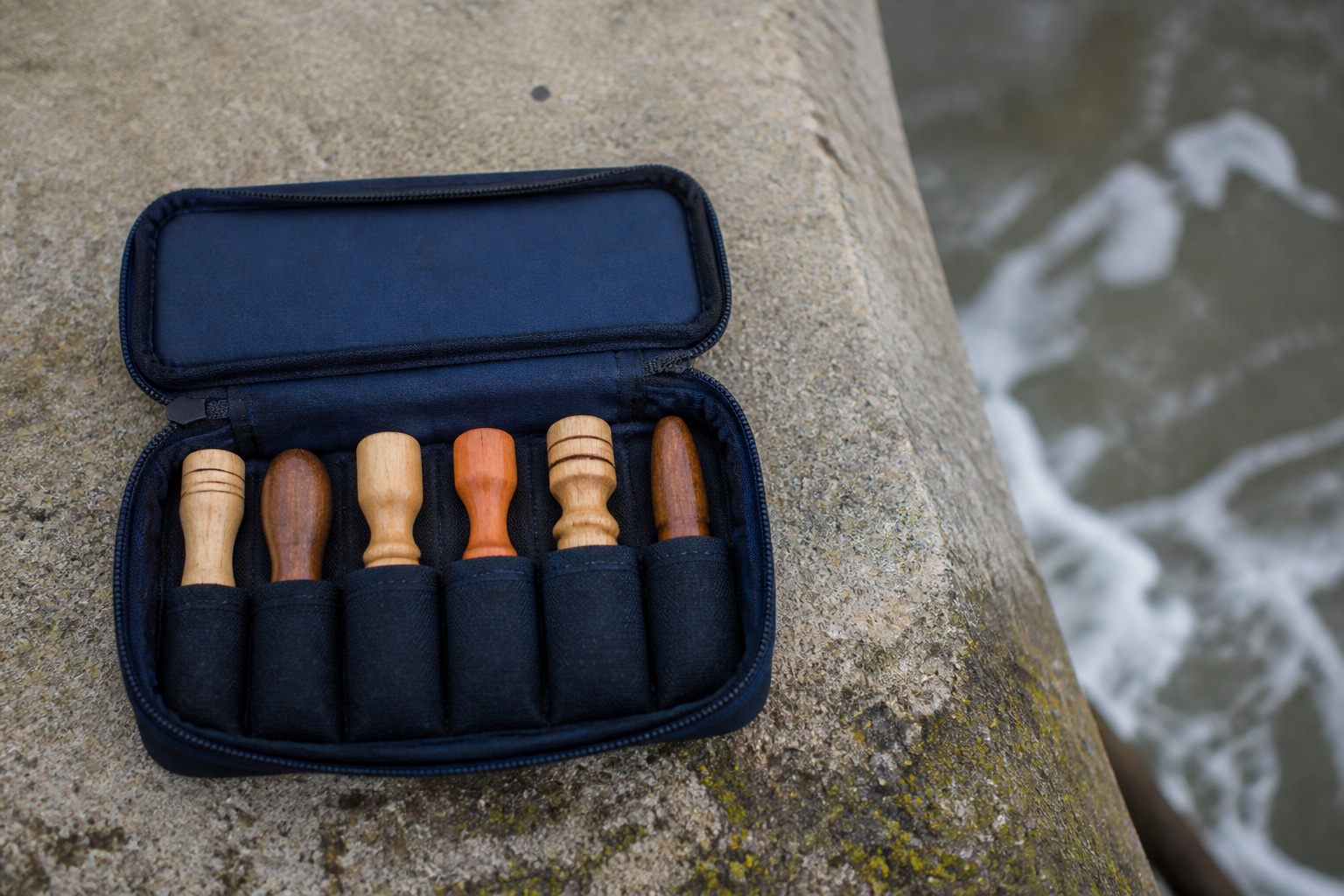 Open surf bag with tube slots holding six wooden plugs of different shapes, seen from above on a jetty wall